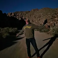Customer-uploaded image. Click to see more details. The image represents: Person walking on sandy ground at night under starry sky with rocky desert landscape in background