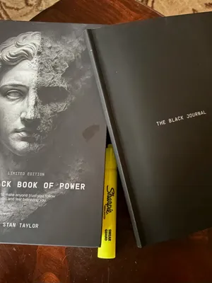 Customer-uploaded image. Click to see more details. The image represents: Two black books on a wooden surface - one titled "The Black Book of Power" by Stan Taylor with a classical statue image on the cover, and another titled "The Black Journal" alongside a yellow pen