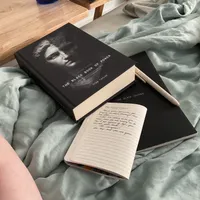 Customer-uploaded image. Click to see more details. The image represents: A book titled "The Black Book of Power" with a portrait on the cover lies on light blue bedding next to an open journal with handwritten text and a pen.