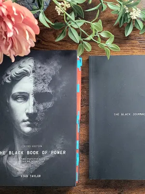 Customer-uploaded image. Click to see more details. The image represents: Two black books on a wooden surface with flowers - one titled "The Black Book of Power" by Stan Taylor with a classical statue image, and another titled "The Black Journal"