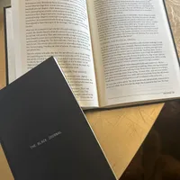 Customer-uploaded image. Click to see more details. The image represents: An open book displaying text pages sits next to a black journal titled "THE BLACK JOURNAL" on a wooden surface