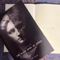 Customer-uploaded image. Click to see more details. The image represents: A black and white book cover titled "The Black Book of Power" by Stan Taylor, featuring a classical statue portrait, placed on top of other books on a textured surface.