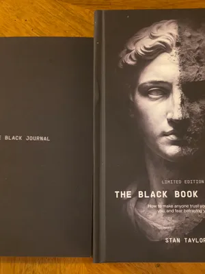 Customer-uploaded image. Click to see more details. The image represents: Two black books on a wooden surface - one titled "The Black Journal" and another titled "The Black Book of Power" by Stan Taylor with a classical statue face on the cover